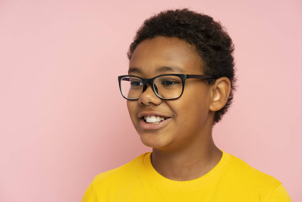 Retrato de niño de escuela afroamericano guapo sonriente con gafas con estilo y camiseta amarilla mirando hacia otro lado aislado sobre fondo rosa. Concepto educativo - Foto, imagen