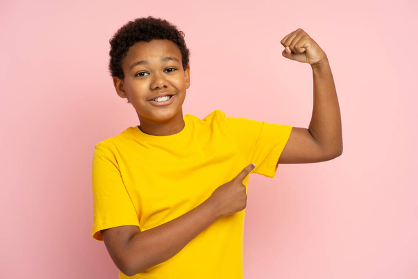 Portret uśmiechniętej, pewnej siebie, silnej, afroamerykańskiej nastolatki wskazującej palcem na bicepsy noszące luźną koszulkę odizolowaną na różowym tle. Atrakcyjne dziecko pozowanie, patrząc na kamerę w studio - Zdjęcie, obraz