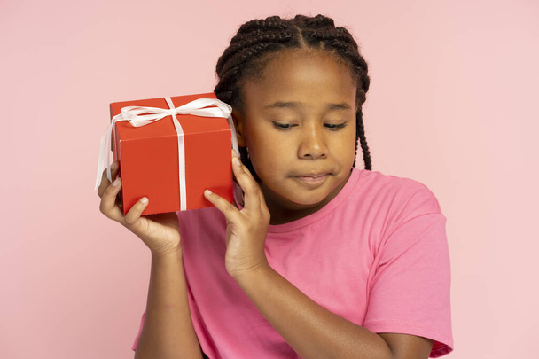 Portrait of attractive little Nigerian little girl holding red gift box, put it to her ear, listening isolated on pink background. Cute child received gift for New Year. Celebration concept - Фото, изображение