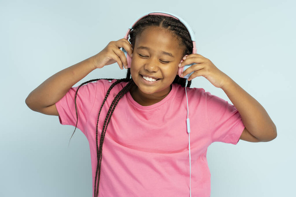 Retrato de chica afroamericana atractiva sonriente con ojos cerrados escuchando música en auriculares aislados sobre fondo azul, espacio para copiar. Linda sonriente pequeña hembra disfrutando - Foto, Imagen