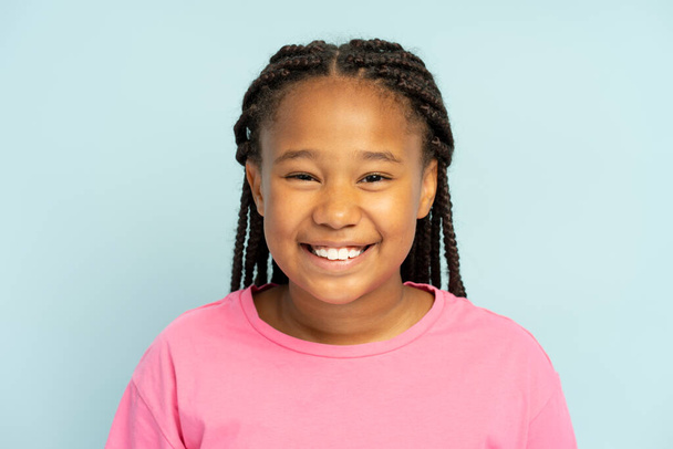 Portrait of cute smiling African American girl wearing stylish hairstyle, dreadlocks, pigtails wearing casual pink t shirt standing isolated on blue background, looking at camera, closeup - Fotografie, Obrázek