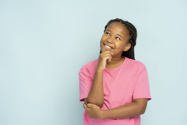 Beautiful smiling Nigerian girl with stylish hairstyle in casual clothes standing looking away, thinking isolated on blue background. Positive child posing. Childhood concept - Фото, изображение