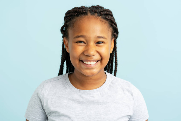 Portrait of cute smiling African American girl wearing stylish hairstyle, dreadlocks, pigtails wearing casual gray t shirt standing isolated on blue background, looking at camera, closeup - 写真・画像