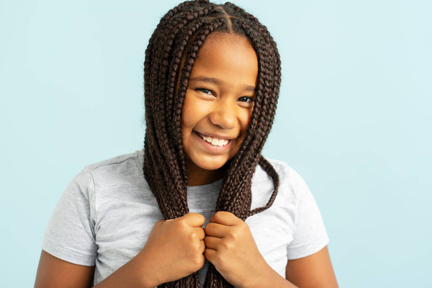 Portrait of smiling positive beautiful African American girl with stylish pigtails, hairstyle holding hair looking in mirror isolated on blue background. Hair care concept - Photo, image