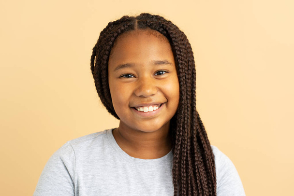 Portrait of beautiful smiling African American girl with stylish hairstyle, pigtails in gray t shirt looking in mirror isolated on beige background. Female child posing in studio. Hair care concept - Fotoğraf, Görsel