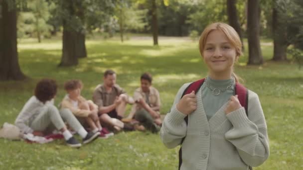 Mittleres Porträt einer hübschen kaukasischen Schülerin mit Rucksack, die an sonnigen Tagen im Freien im öffentlichen Park in die Kamera lächelt, während ihre vielfältigen Klassenkameraden und ihr männlicher Lehrer im Hintergrund auf Gras sitzen - Filmmaterial, Video