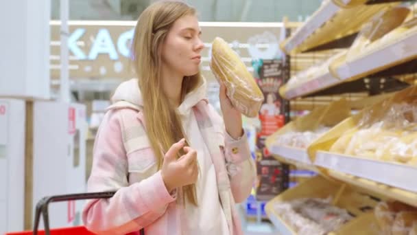 Portrait of blonde young woman buying fresh wheat bread, loaf and rye bread smelling it in supermarket. Bread in plastic bag packaged. Shopping food in grocery hypermarket. - Footage, Video