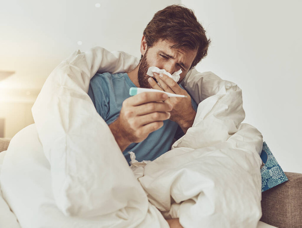Thermometer, sneeze and sick man on sofa with headache, fever or influenza crisis at home. Anxiety, high temperature and male with toilet paper, worry or blowing nose in living room with covid fear. - Fotoğraf, Görsel