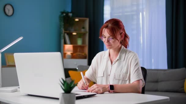 trabajo en casa, mujer joven en gafas trabaja en casa en la computadora hace notas en el cuaderno mientras está sentado en una mesa en la habitación - Imágenes, Vídeo