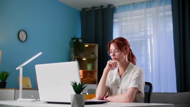 freiberufliche, nachdenkliche junge Frau mit Brille arbeitet am Computer und macht sich Notizen in einem Notizbuch, während sie zu Hause am Tisch sitzt - Filmmaterial, Video