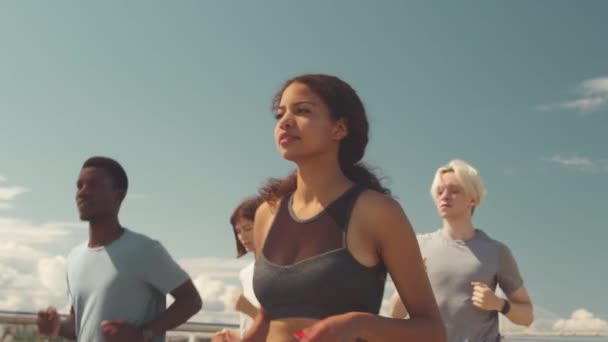 Mittleres Zeitlupentempo von vier jungen Sportlerinnen und Sportlern beim sommerlichen Joggen entlang der Brücke - Filmmaterial, Video