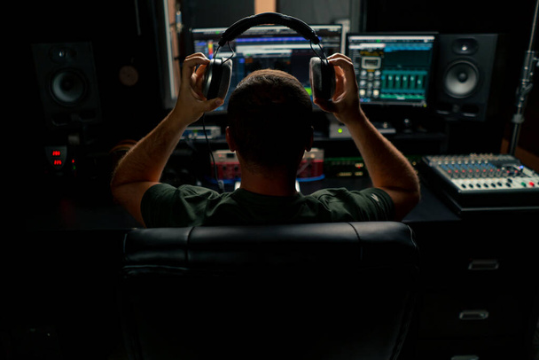 男性のサウンドエンジニアがカメラに背中が付いているミキシングデスクに座ってヘッドフォンを置きます - 写真・画像