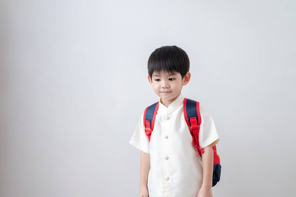 Aasialainen poika yllään perinteisiä thaimaalaisia vaatteita, seisoo koulun laukku. valmistautuminen kouluun valkoisella pohjalla - Valokuva, kuva