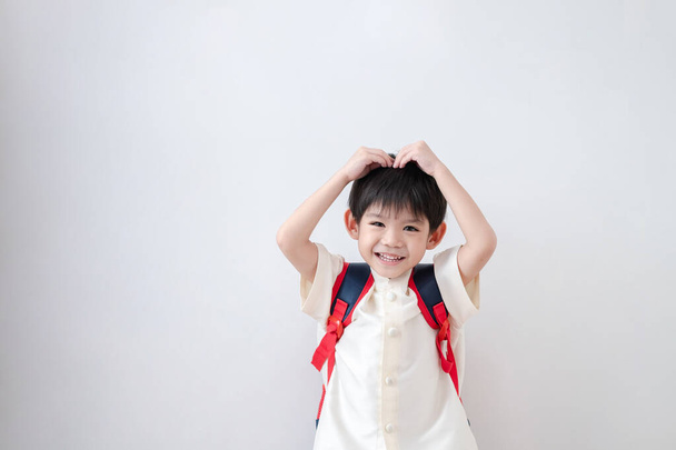Aasialainen poika yllään perinteisiä thaimaalaisia vaatteita, seisoo koulun laukku. Tee sydämen ele hänen päänsä valmistautuu menemään kouluun valkoisella taustalla - Valokuva, kuva