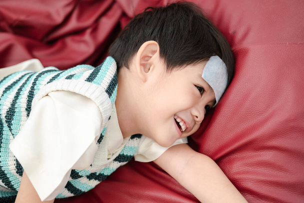Azjatycki chłopiec Leżący chory z gorączką na czole, ale nadal uśmiechnięty. - Zdjęcie, obraz