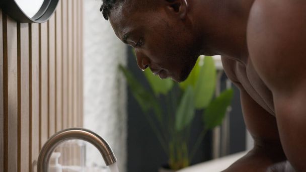 Kylpy aamu rutiini Afrikkalainen Amerikkalainen mies pesu kasvot kaatamalla kylmää vettä kylpyhuoneessa pesuallas etninen mies pese puhdas kasvojen ihonhoito hygienia kosteuttava virkistävä anti akne menettely hoito - Valokuva, kuva