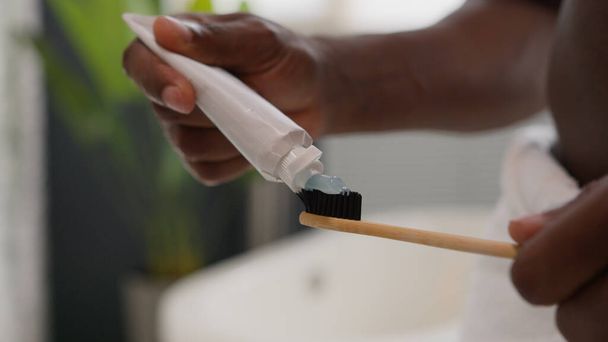 Zamknij ręce wyciskając białą pastę do zębów z tubki na szczoteczce do zębów zabieg higieny jamy ustnej rano zapobiec próchnicy nieznany Afroamerykanin przygotować zęby mycia w łazience - Zdjęcie, obraz