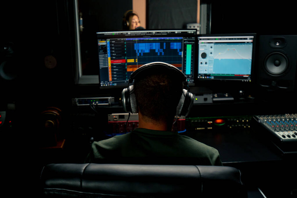 ヘッドフォンを身に着けている男性のサウンドエンジニアが,カメラの背中にあるミキシングデスクに座って,コンピュータモニターのサウンドトラックを見ます. - 写真・画像