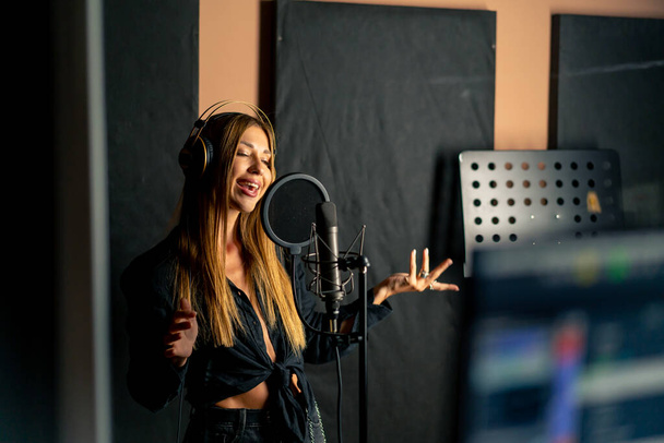 piosenkarka śpiewa w studiu nagraniowym ze słuchawkami przed mikrofonem, aby stworzyć nową piosenkę - Zdjęcie, obraz