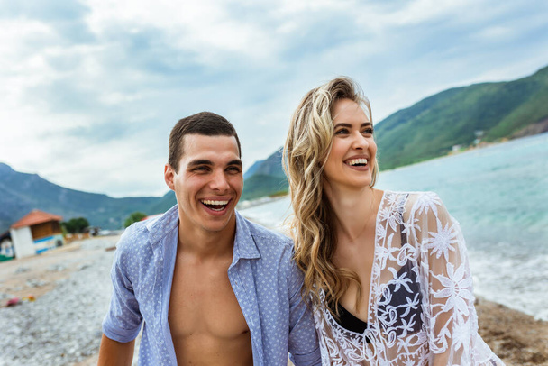 Ευτυχισμένο νεαρό ζευγάρι να γελάει και να κρατιέται χέρι-χέρι στην παραλία - Φωτογραφία, εικόνα