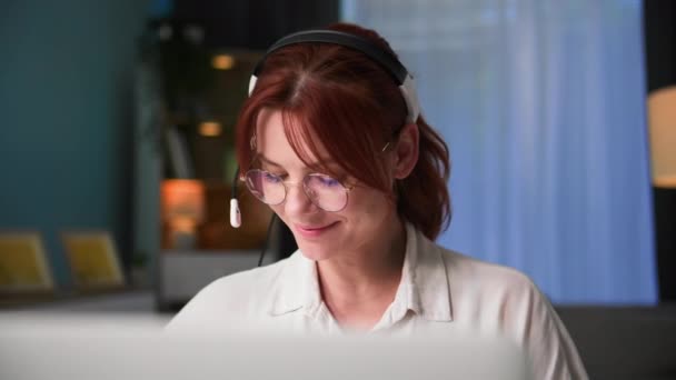 werk op afstand, jonge vrouw in bril werkt op een computer en praat via video link met behulp van headset terwijl zitten aan een tafel in een kamer - Video