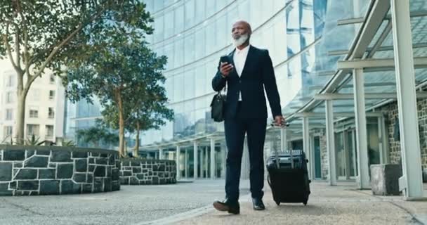 Phone, suitcase and businessman walking in city for corporate work trip flight by airport. Travel, luggage and professional African executive ceo networking on cellphone by building in urban town - Footage, Video