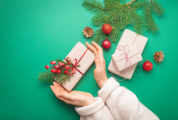 Noël ou Nouvel An célébration papier vert fond festif avec les mains féminines tenant boîte cadeau enveloppé, sapin de décoration, cônes, baies, boules rouges scintillantes. Espace pour le texte.. - Photo, image