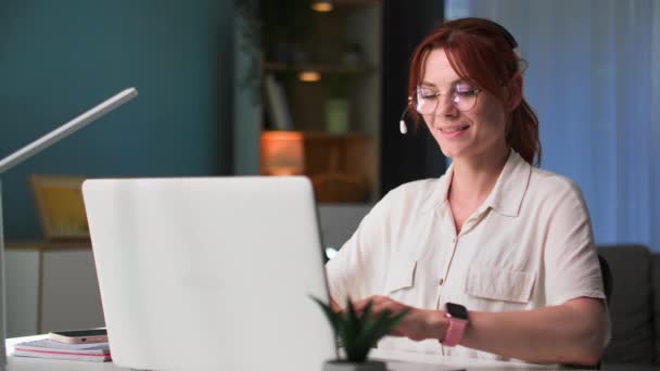 szabadúszó, szemüveges fiatal nő dolgozik a számítógépen, és beszél videokapcsolaton keresztül az ügyféllel egy headset és jegyzetel egy notebook, miközben ül az asztalnál egy szobában - Felvétel, videó