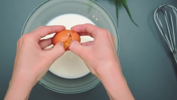 Weibliche Hände brechen ein Ei in eine tiefe Glasschale mit Milch. Draufsicht auf den Küchentisch. Kochen in der Küche - Filmmaterial, Video