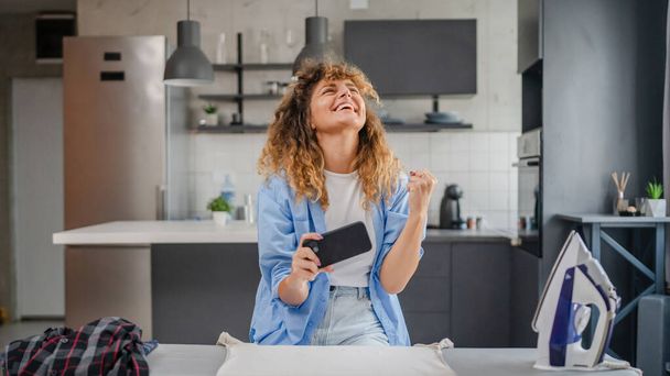 One adult young caucasian woman play video games on smartphone at home have fun while take a brake from housework real person leisure concept copy space - Photo, Image