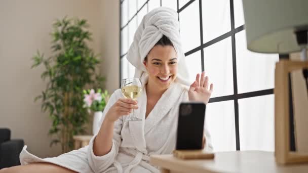 Young beautiful hispanic woman wearing bathrobe drinking champagne having video call at bedroom - Footage, Video