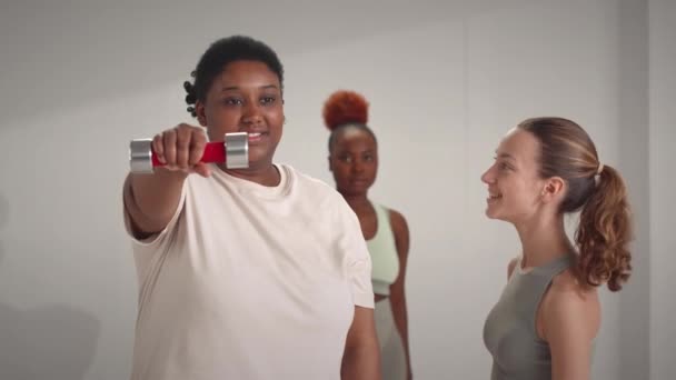 Medium shot van jonge blanke vrouwelijke fitnesscoach tonen hoe te doen halter front verhogen oefening aan twee jonge zwarte vrouwen tijdens de groep fitness training binnen - Video