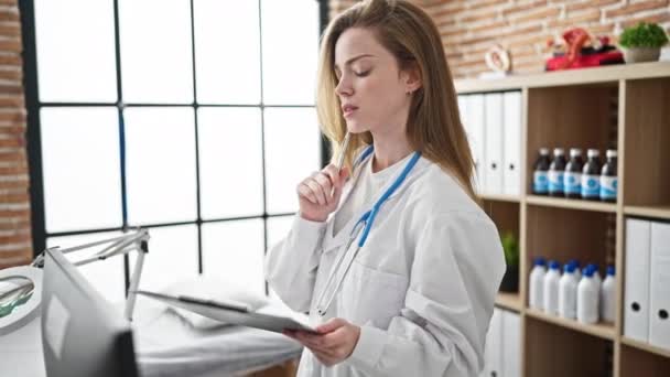 Νεαρή ξανθιά γυναίκα γιατρός έγγραφο γραφής στο πρόχειρο στην κλινική - Πλάνα, βίντεο