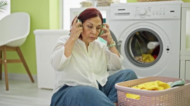 Жінка середнього віку слухає музику прання одягу в пральні - Кадри, відео
