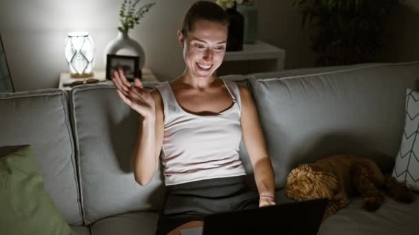Молодая белая женщина с собакой делает видеозвонок, используя ноутбук, сидя дома на диване - Кадры, видео