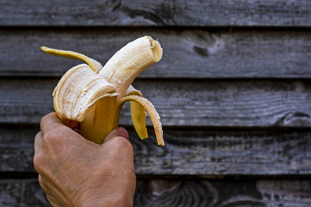 držet pokousaný kus oloupaného zralého banánu v ruce na pozadí dřevěného plotu - Fotografie, Obrázek