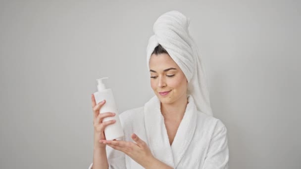Young beautiful hispanic woman wearing bathrobe holding lotion bottle over isolated white background - Footage, Video