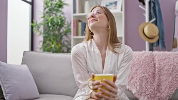 Mujer rubia joven bebiendo café sentado en el sofá sonriendo en casa - Metraje, vídeo