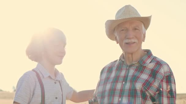 Горизонтальный выстрел. Веселый пожилой мужчина обнимается с парнем. Счастливого дедушки и внука, стоящих на поле. Мужчина в клетчатой рубашке и соломенной шляпе, обнимающий ребенка. Сельское хозяйство или выращивание. - Кадры, видео