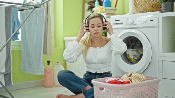 Młoda blondynka słuchająca muzyki pranie ubrań w pralni - Materiał filmowy, wideo