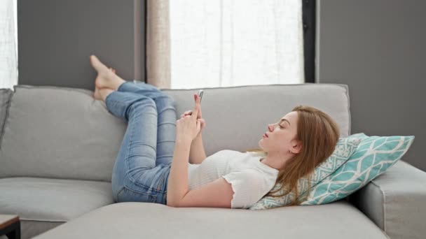 Nuori blondi nainen käyttää älypuhelinta makaa sohvalla kotona - Materiaali, video