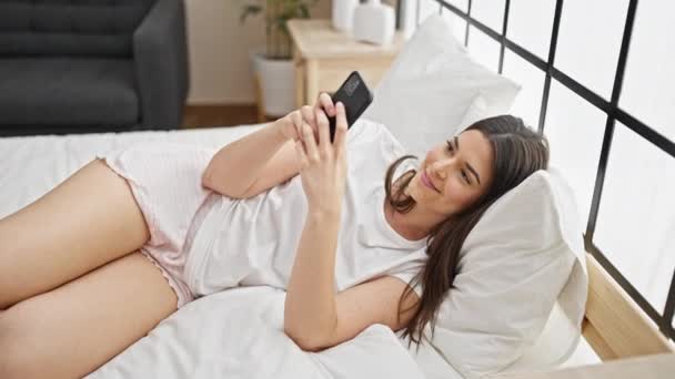 Νεαρή όμορφη ισπανόφωνη γυναίκα που χρησιμοποιεί smartphone ξαπλωμένη στο κρεβάτι στο υπνοδωμάτιο - Πλάνα, βίντεο
