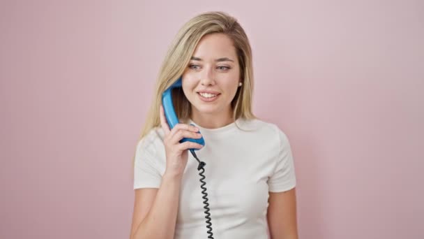 Молодая блондинка уверенно улыбается, разговаривая по телефону с победителем жеста на изолированном розовом фоне - Кадры, видео