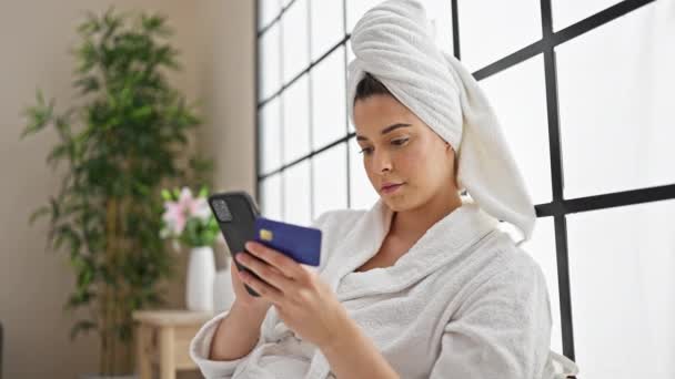 Young beautiful hispanic woman wearing bathrobe shopping with smartphone and credit card at bedroom - Footage, Video