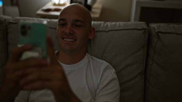 自宅で笑顔でソファーに座ってスマートフォンを使用する若いヒスパニック男性 - 映像、動画