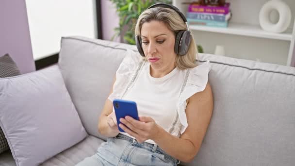 Mladá blondýna žena pomocí smartphone nosí sluchátka sedí na pohovce doma - Záběry, video