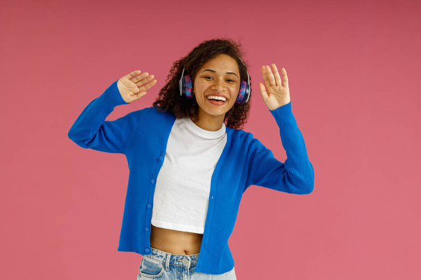 Smiling woman in headphones dancing with raised hands standing on pink studio background - 写真・画像