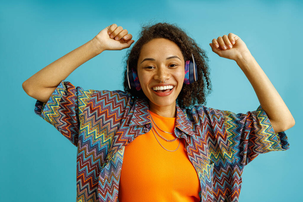 Smiling woman in casual clothes dancing with raised hands standing over blue studio background - Foto, Imagen