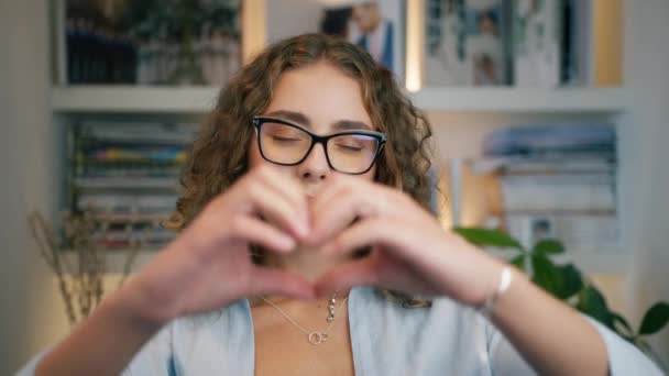 Portrait of attractive female student with curly brunette hair making heart shape with hands and looking to camera in modern apartment. Smiling model in stylish glasses with black modern frame 4K - Footage, Video