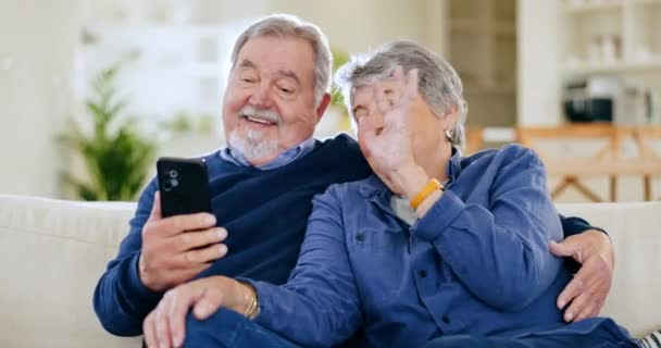 Ηλικιωμένοι, κύμα και ζευγάρι με τηλέφωνο για βιντεοκλήση στον καναπέ με αγκαλιά, επικοινωνία και ευτυχία στο σαλόνι. Senior, άνδρας και γυναίκα με smartphone στον καναπέ με χαιρετισμό σε απευθείας σύνδεση για συνομιλία. - Πλάνα, βίντεο